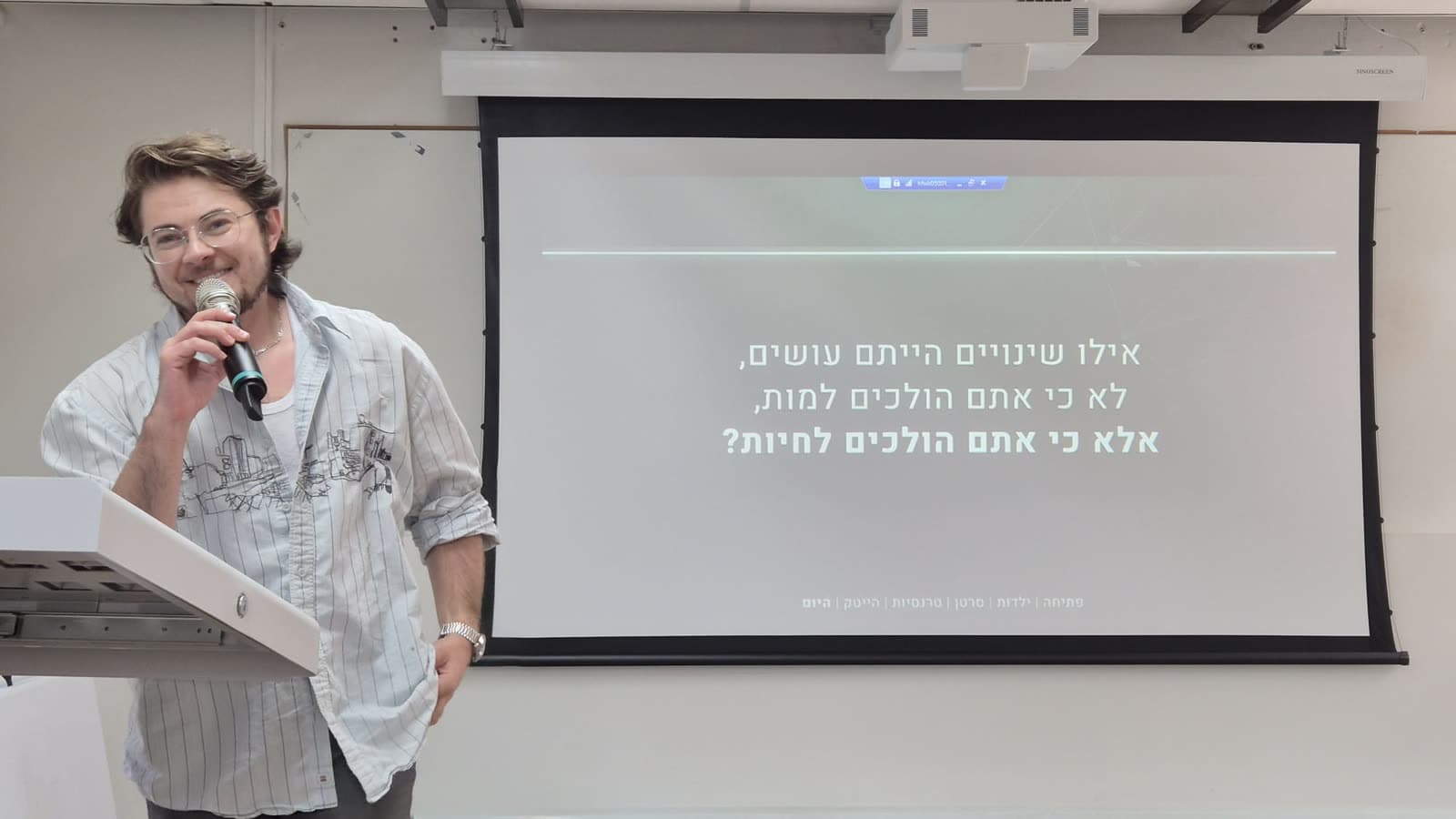 ג'סי ברנדט מחייך עם מיקרופון ליד שקופית מתוך ההרצאה