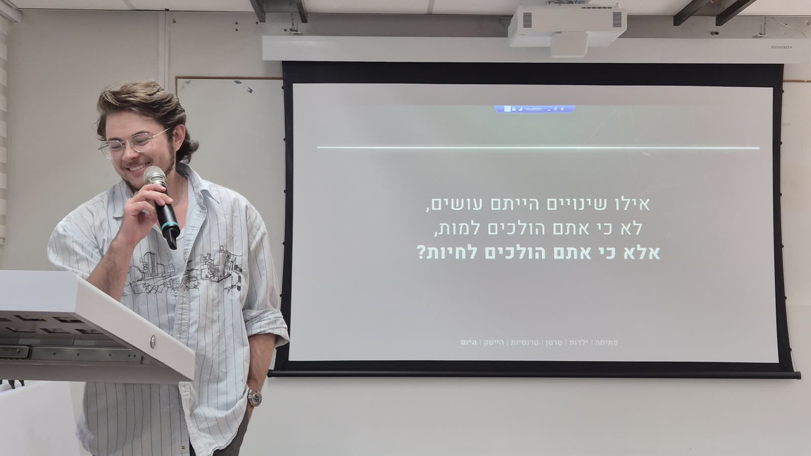 ג'סי ברנדט מחייך ומביט מטה, המיקרופון ביד, ליד שקופית מתוך ההרצאה